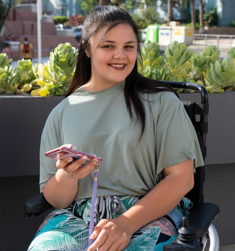 A young woman in a wheelchair smiling and holding a smartphone, representing independence and accessibility through NDIS support in Sydney.
