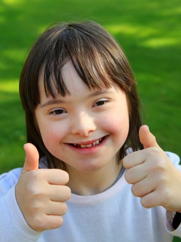 A young girl with Down syndrome smiling and giving two thumbs up, symbolizing positivity and inclusion through NDIS support in Sydney.