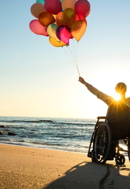 A person in a wheelchair holding colorful balloons and enjoying the sunset at the beach, symbolizing freedom and positivity through NDIS support.